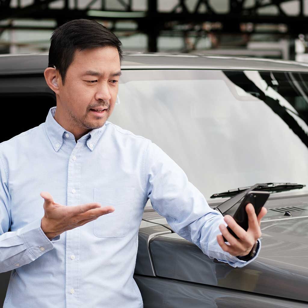 An Asian man is standing outside next to a car while video chatting.