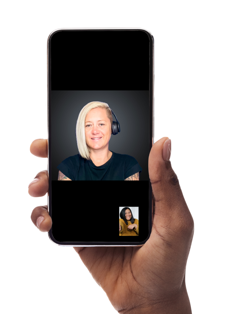 A Polynesian right hand is holding a black smartphone device. On the screen, there is a visible Auslan interpreter. The interpreter is a Caucasian woman with short blonde hair, wearing a black headset and a black shirt. She has tattoos on both arms and is smiling in front of a grey background. At the lower right corner of the screen, there is a small image of a Polynesian woman with long black hair, wearing a mustard-coloured jumper. She is also holding the smartphone in her right hand.